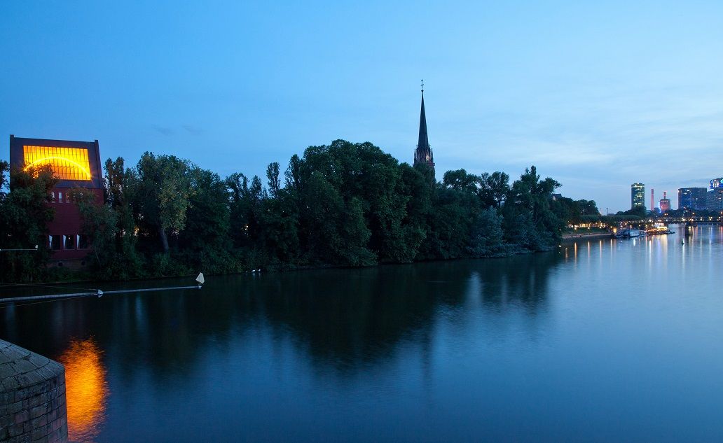 Main in den Abendstunden mit dem Portikus auf der linken Seite mit Lichtinstallation im Dach und Kirchturm mittig im Bild
