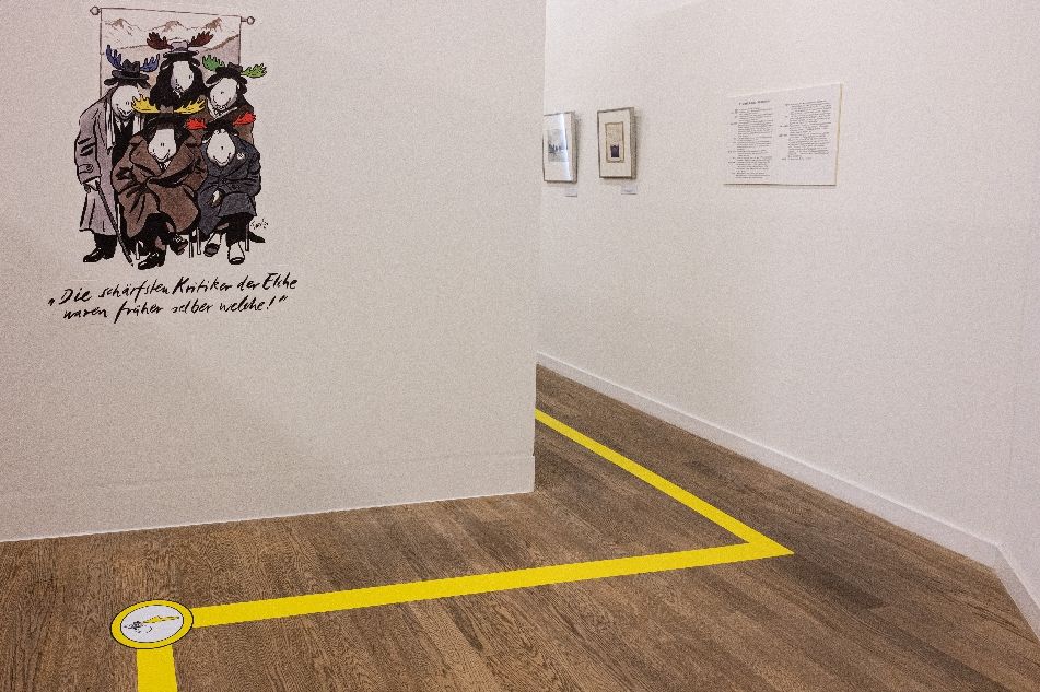 Blick auf eine Karikatur in der Ausstellung mit Elchen in Mänteln mit der Bildunterschrift "Die schärfsten Kritiker der Elche waren früher selber welche."
